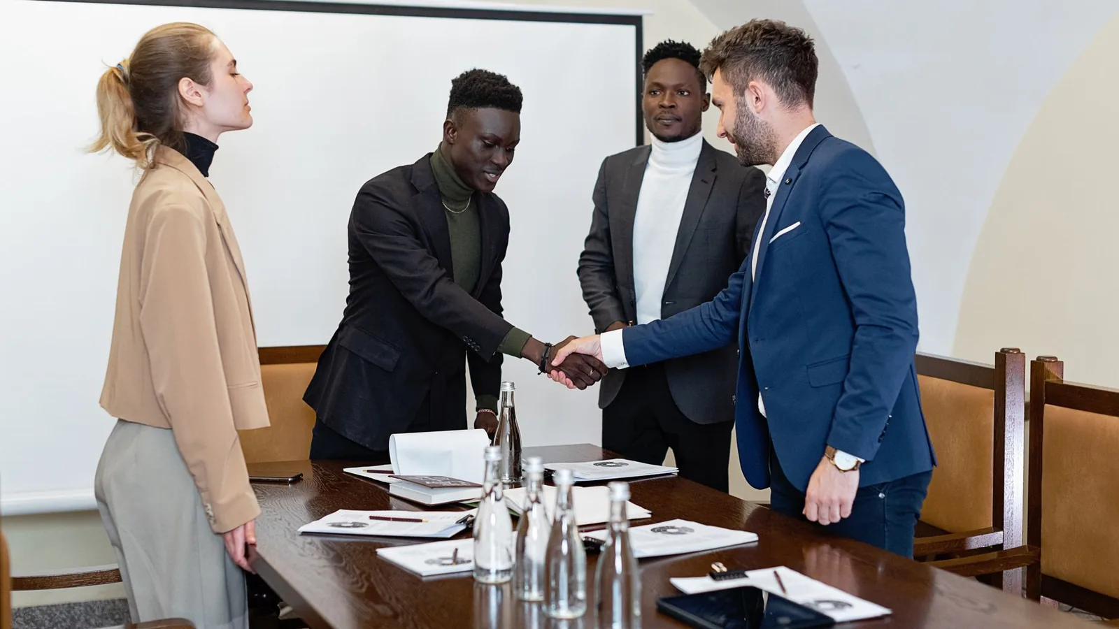 Two business executives shaking hands over a boardroom table with colleagues watching, representing a major business deal