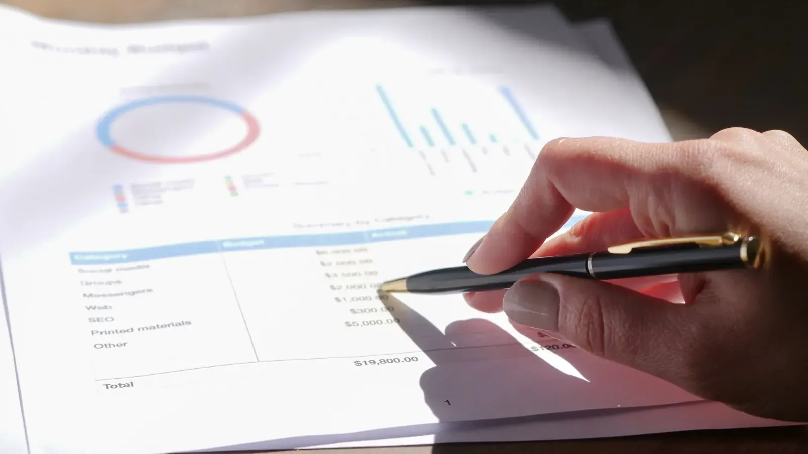 A hand holding a pen over a budget report with a chart, pointing at a line item during a reconciliation check