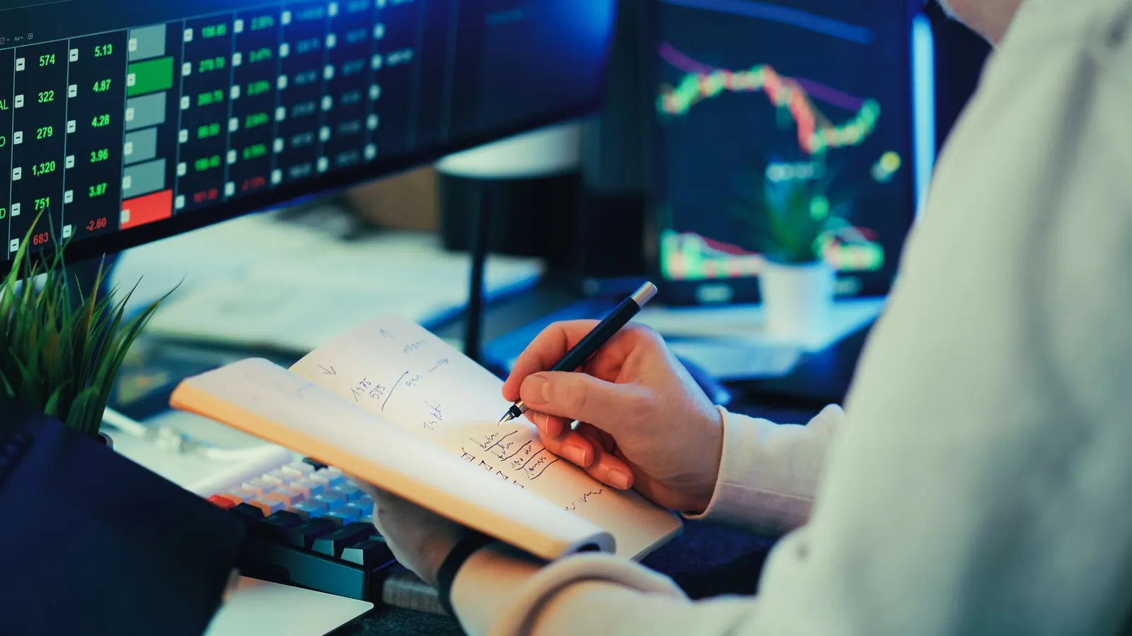 A hand writing notes in a notebook in front of monitoring screens showing data charts, representing the ongoing rhythm of checking reconciliations and reviews