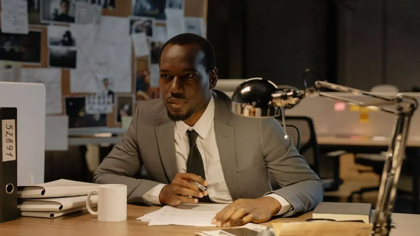 A private investigator reviewing case files at a desk in front of an evidence board