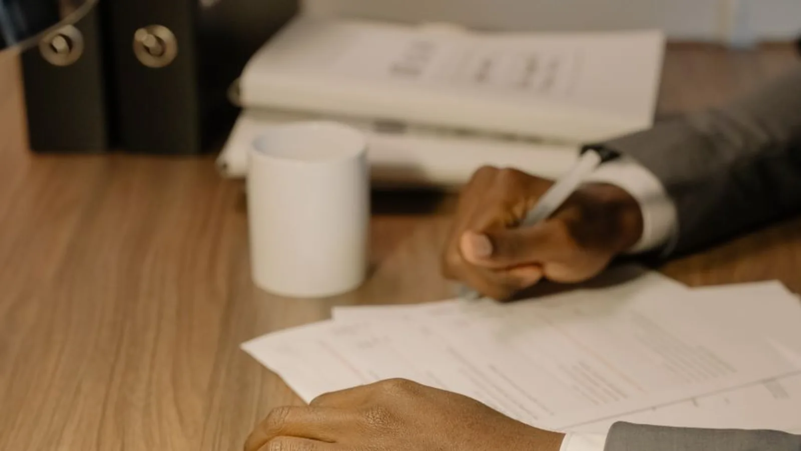 A close-up of hands writing notes over documents at a desk with a coffee mug and folders, representing a documented investigation report