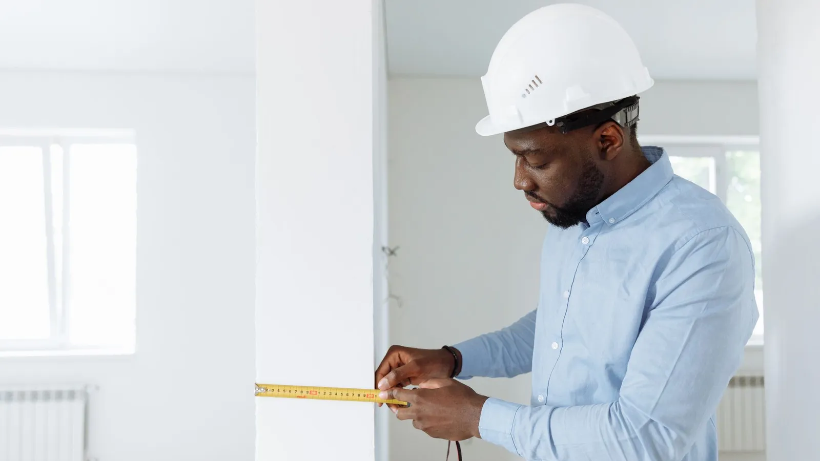A man in a hard hat measuring a wall with a tape measure inside a property, representing the on-site inspection at the center of a loss adjustment