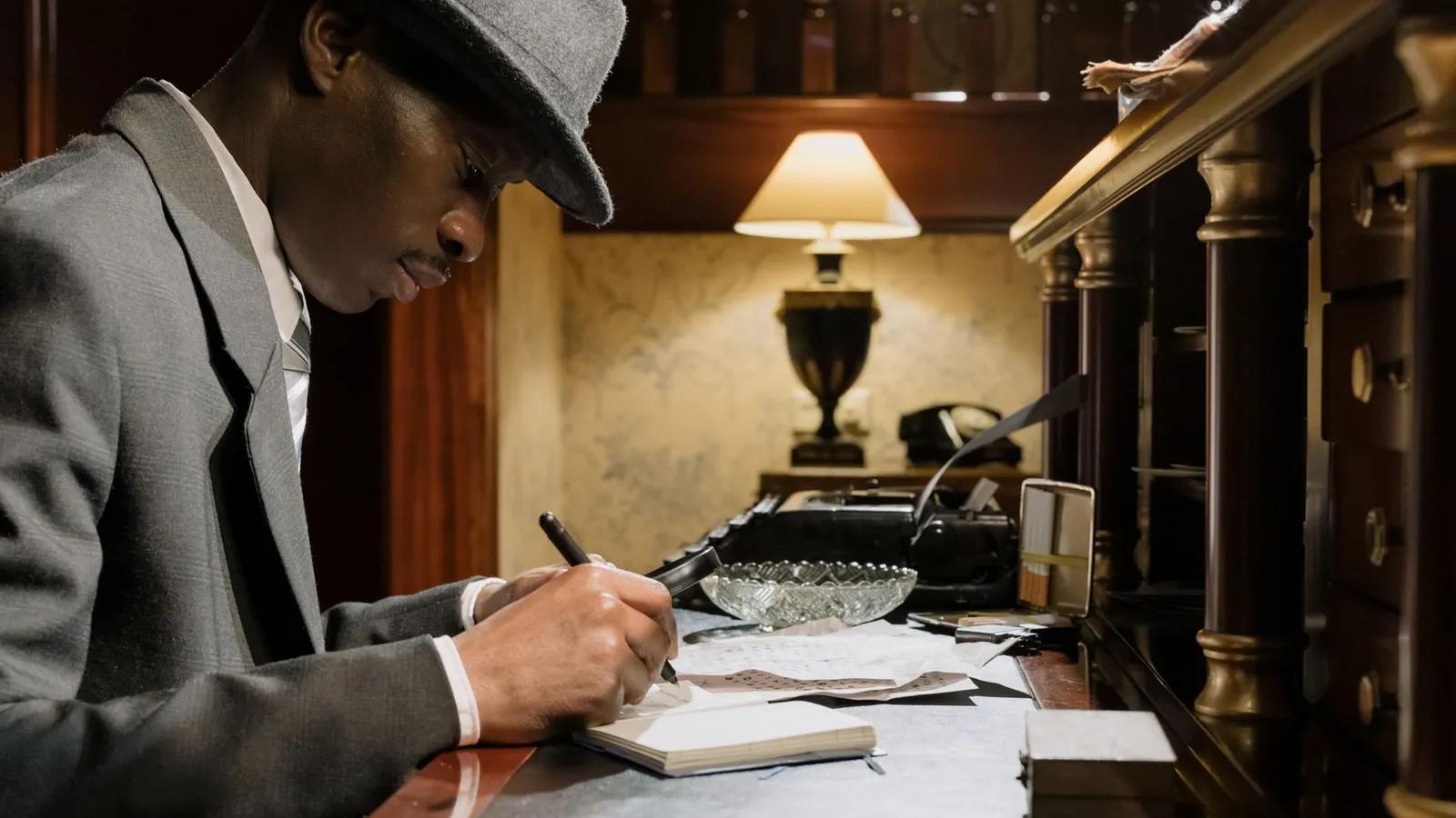 A man in a fedora writing notes at a desk under lamplight, representing the cinematic image most people have of a private investigator