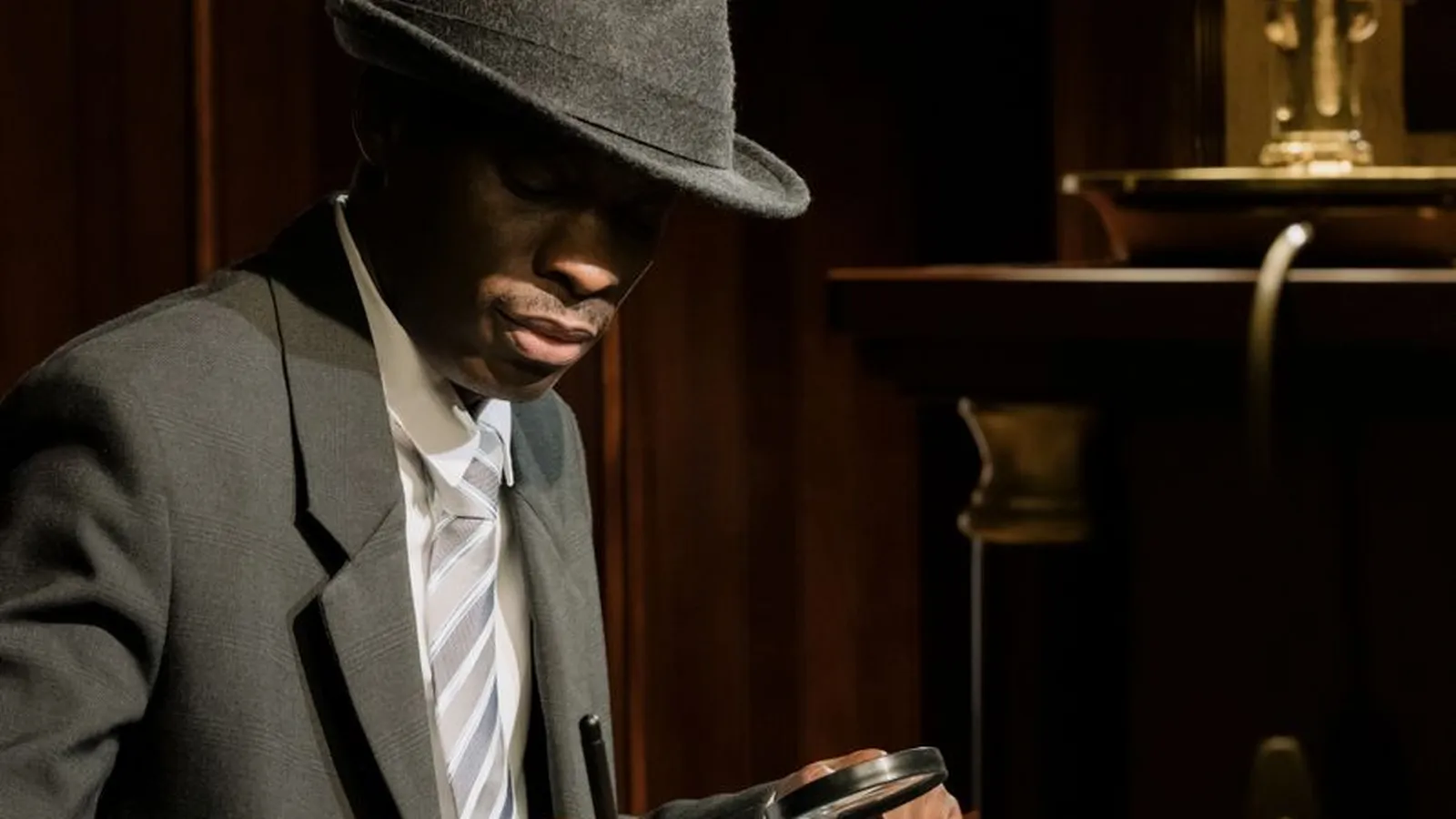 A man in a fedora examining documents with a magnifying glass under lamplight, representing the cinematic cliche the actual work looks nothing like day to day
