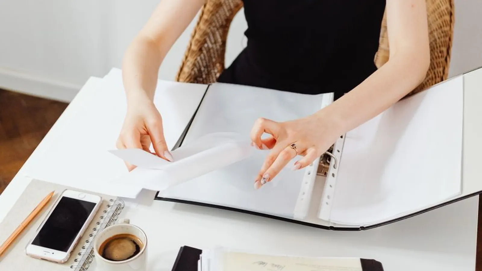 Hands opening a binder full of documents next to a phone and coffee cup on a desk, representing the range of case files a private investigator handles