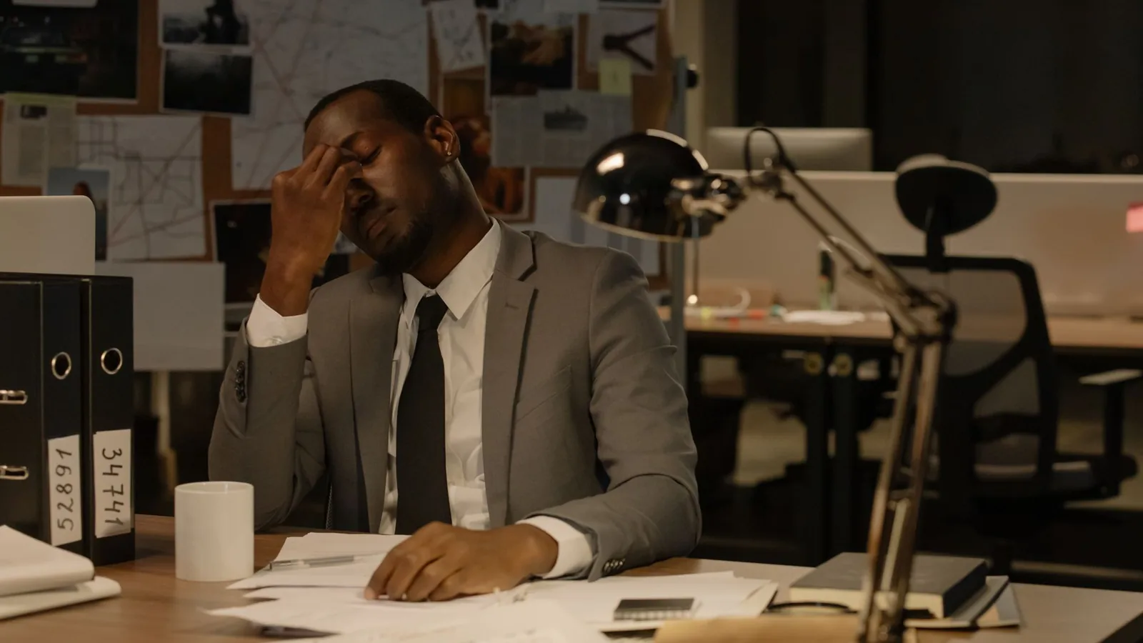 A man at his desk rubbing his temples amid ordinary paperwork, representing that the real work looks like patient documentation, not dramatic movie scenes